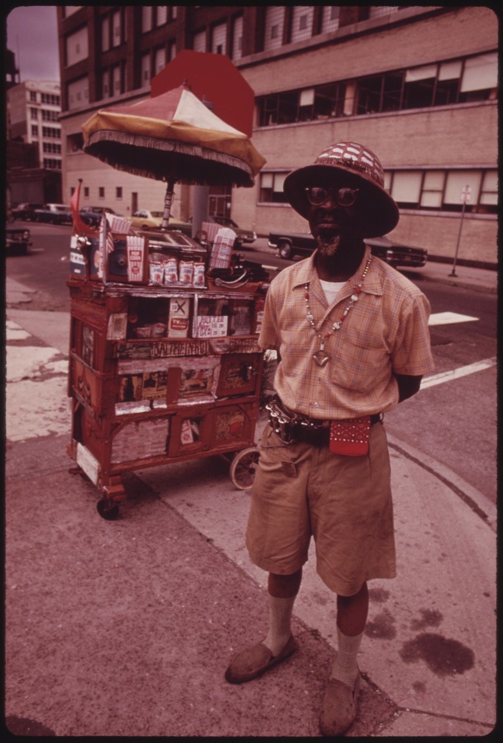 A street vendor stands next to his cart on a city street. The vendor is a Black man with a beard, wearing a patterned hat, sunglasses, a light-colored short-sleeved shirt, khaki shorts, white socks, and brown shoes. The cart is covered in various items for sale, including magazines, newspapers, and other small goods. It has a large red umbrella attached to the top. The background shows a multi-story building with windows and a street with cars parked along the side.