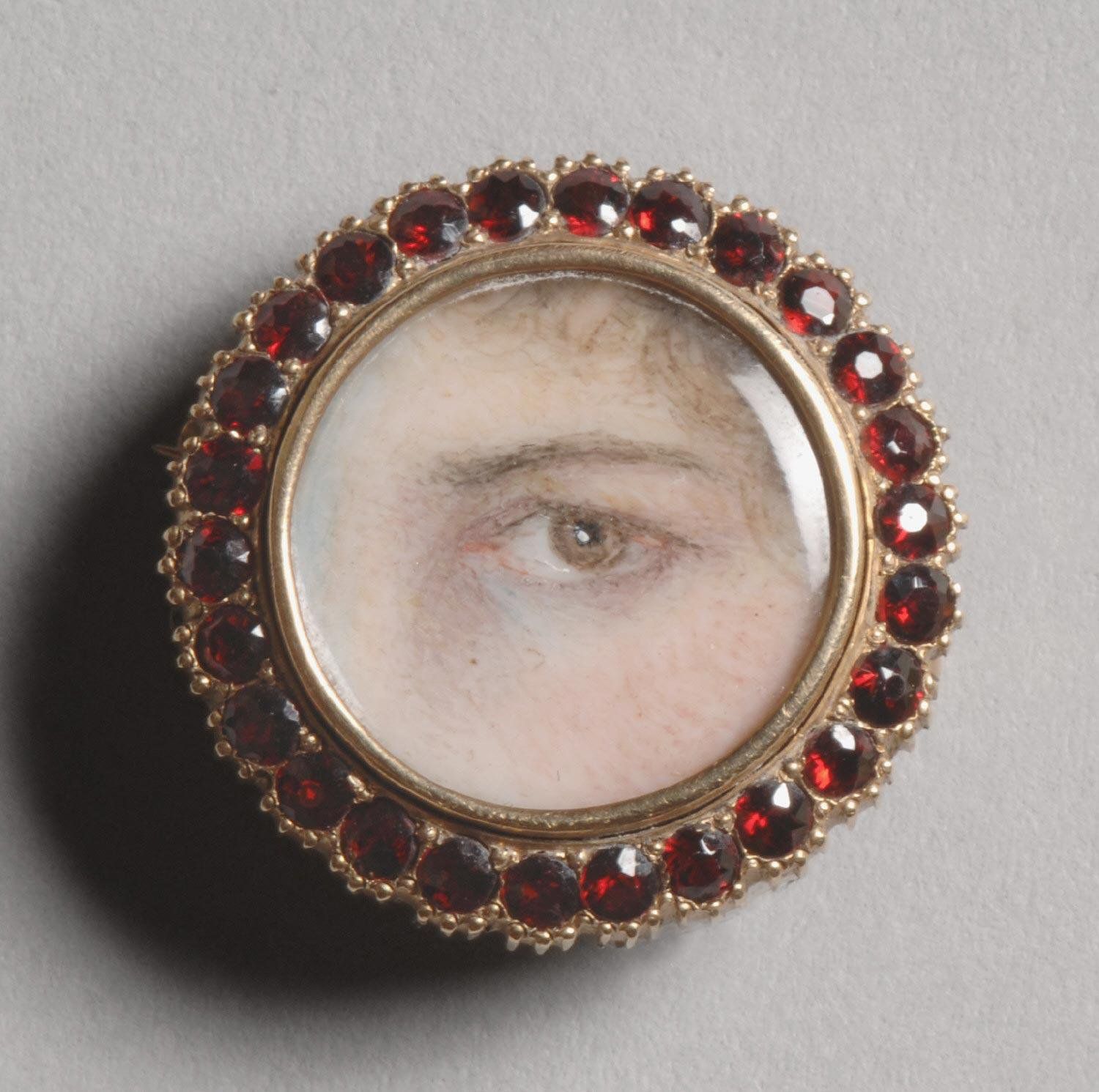 Close-up of a circular brooch featuring a miniature painting of a realistic eye surrounded by a ring of deep red garnets in a gold setting.