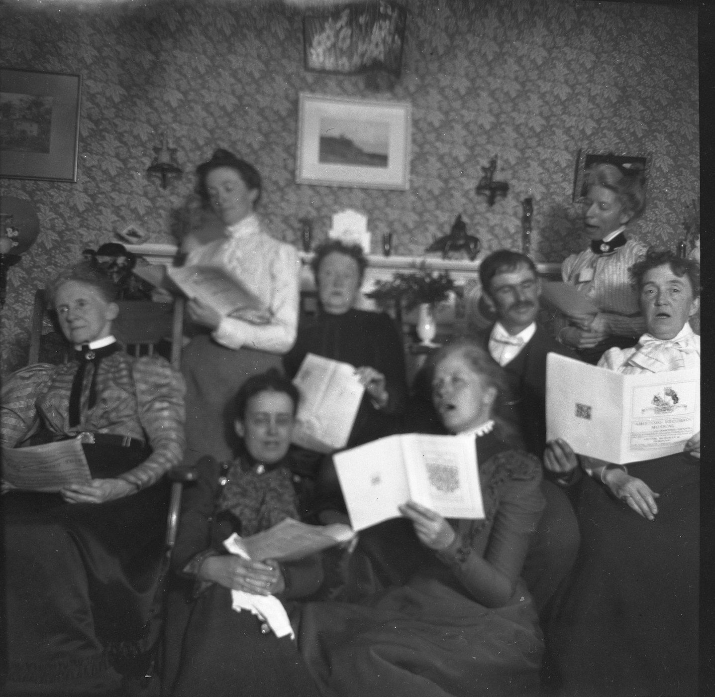 A black and white photograph of a group of people gathered in a room, holding sheet music and appearing to be singing.