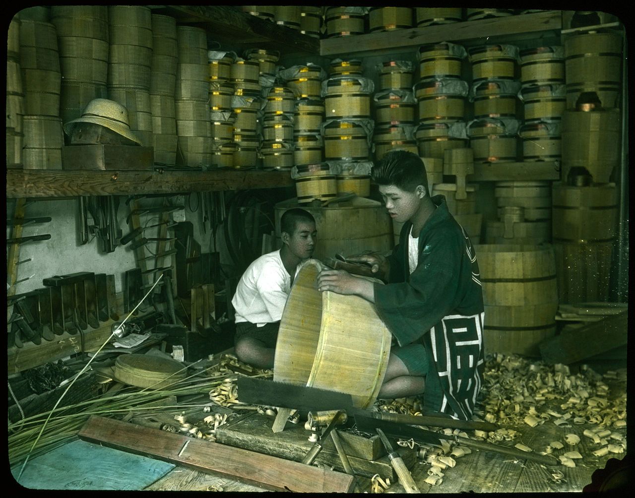 Two young boys working in a traditional barrel-making workshop, surrounded by stacks of finished barrels and piles of wood shavings.