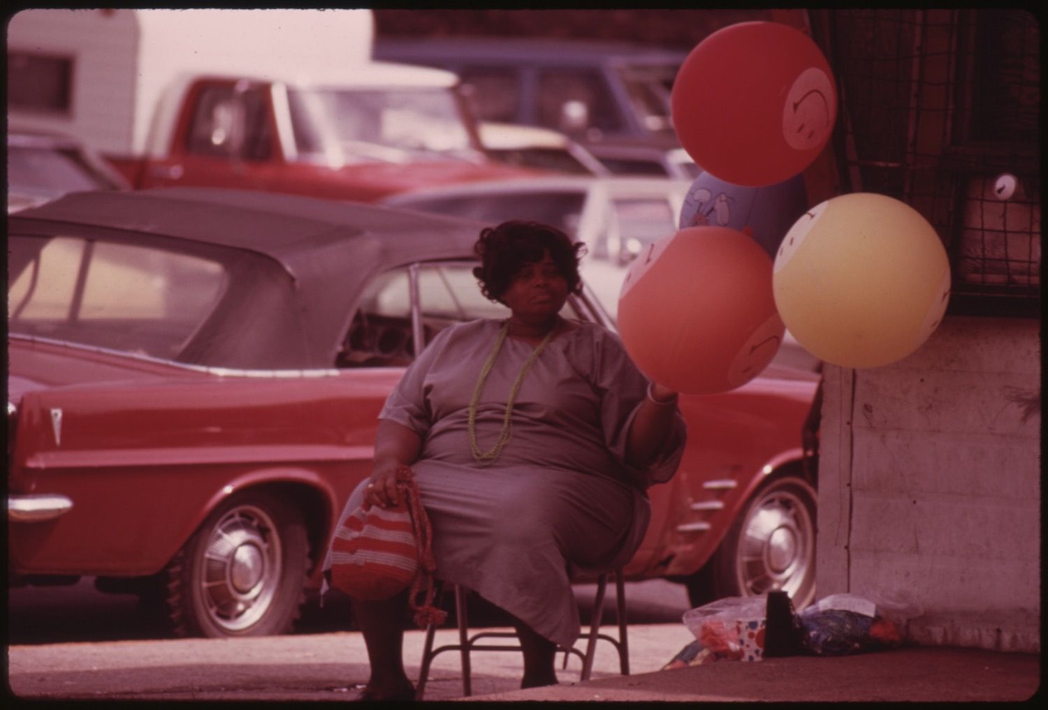 A woman sits on a metal chair surrounded by balloons, with vintage cars parked behind her.