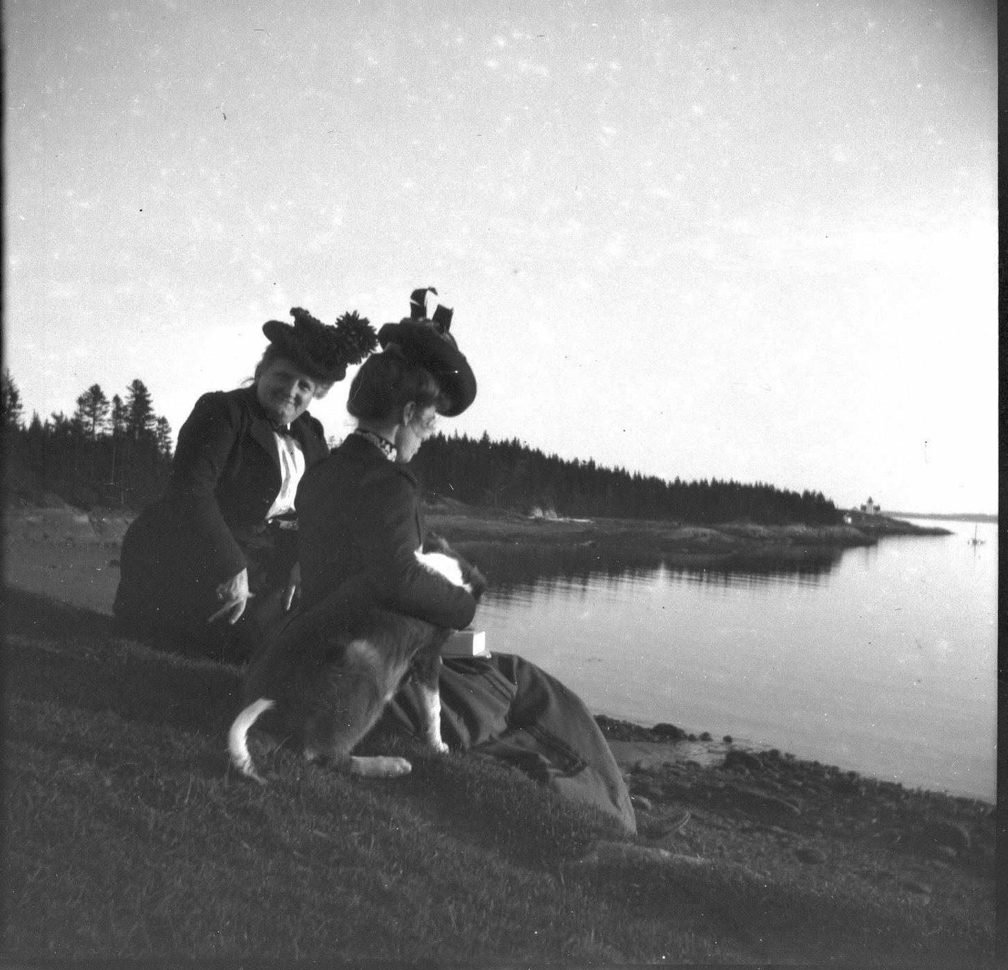 Two women in hats and a dog sit on a grassy bank overlooking a body of water with a forested shoreline in the background.