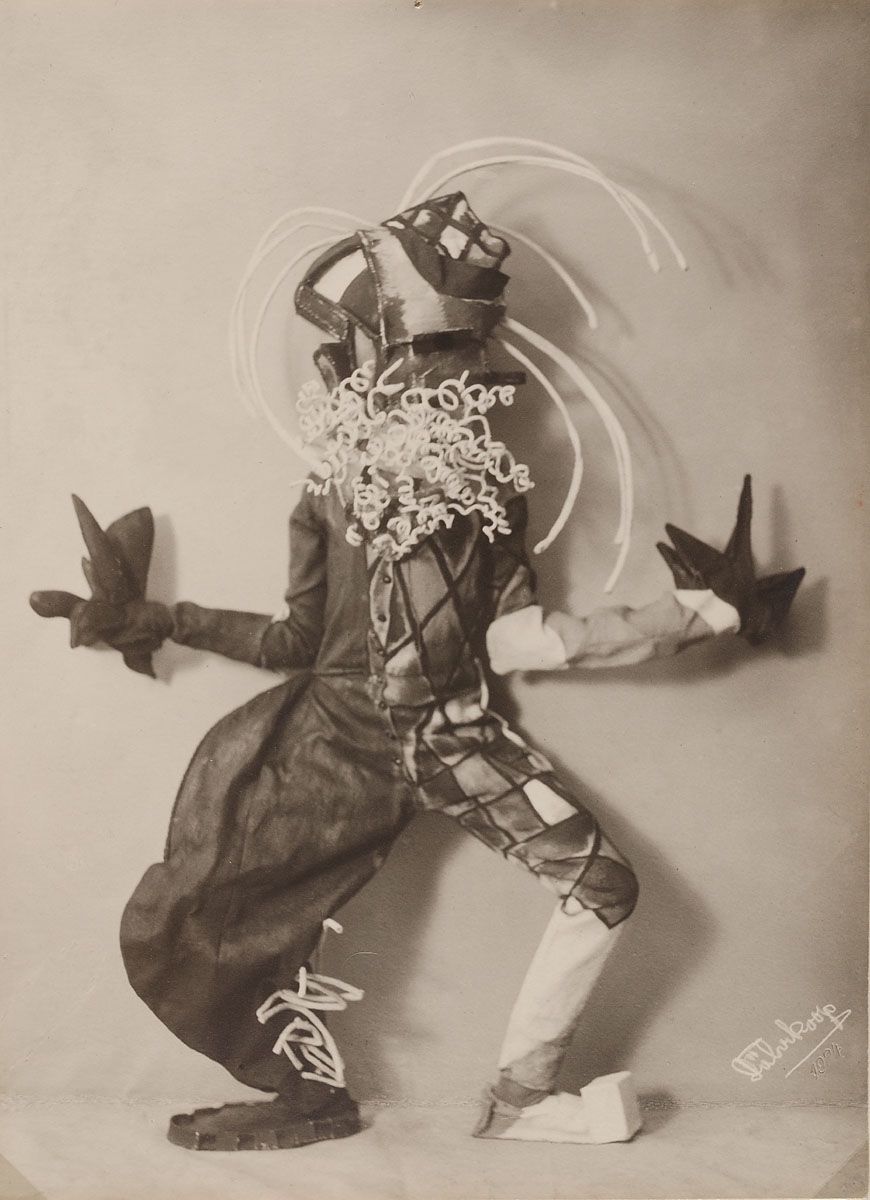 A black and white photograph of a performer in a geometric costume and mask, holding bird-shaped objects, with a floral halo around their head. The photograph is dated 1924.