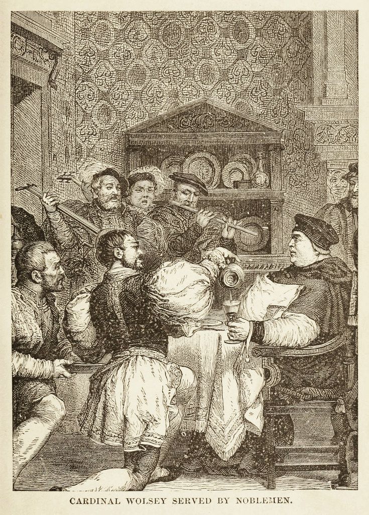 A black and white engraving titled "Cardinal Wolsey Served by Noblemen." The scene depicts Cardinal Wolsey seated at a table, being attended to by numerous noblemen who are serving him food and drink. The room is richly decorated with a fireplace, portraits, and ornate paneling.