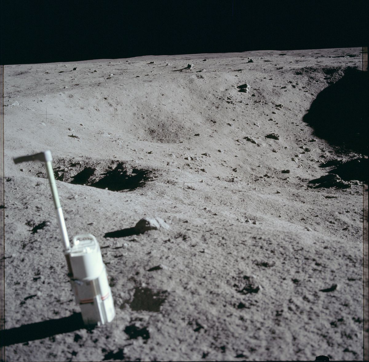 A desolate lunar landscape with a tool planted in the gray soil, a small crater, and a dark rock.