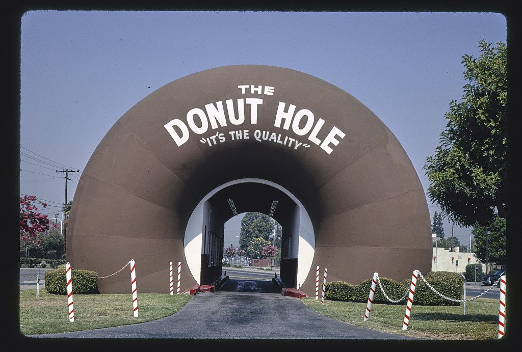 A large, brown, arch-shaped structure resembling a donut hole with the words "THE DONUT HOLE" and "IT'S THE QUALITY" written on it. A road leads through the archway, and red and white striped poles with a rope barrier line the sides. The structure is surrounded by green grass, trees, and buildings under a clear blue sky.