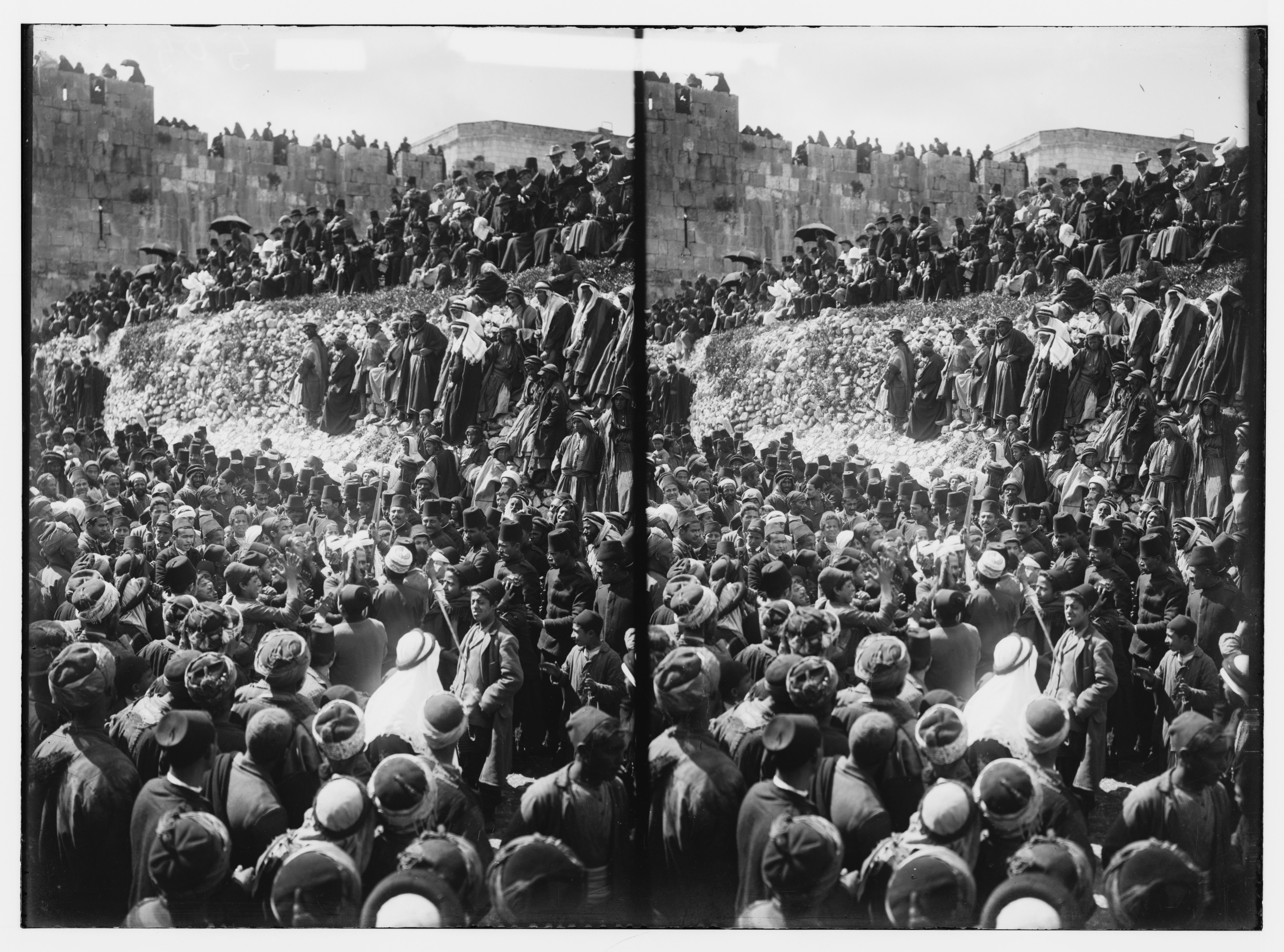 Stereoscopic photograph of a large crowd gathered in front of a massive stone wall.