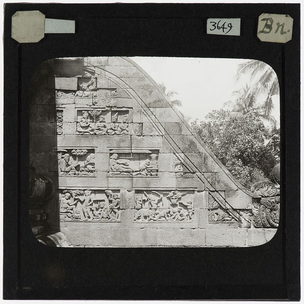 A black and white photograph of a detailed stone carving within an archway, surrounded by vegetation, likely part of an ancient structure.