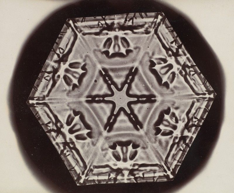 A black and white microscopic photograph of a snowflake with a hexagonal shape and intricate patterns.