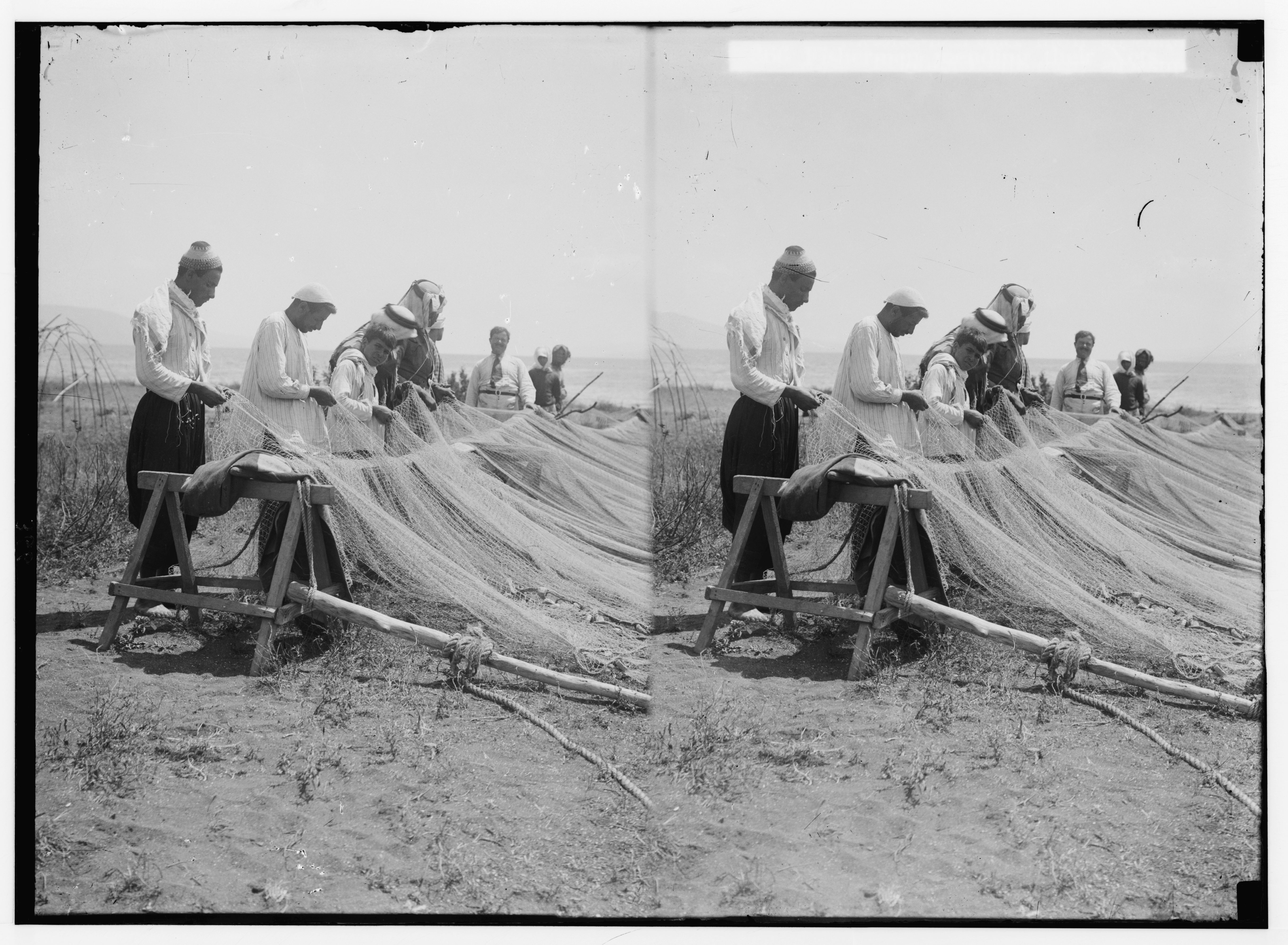 A black and white stereograph showing a group of men working on a large fishing net outdoors.