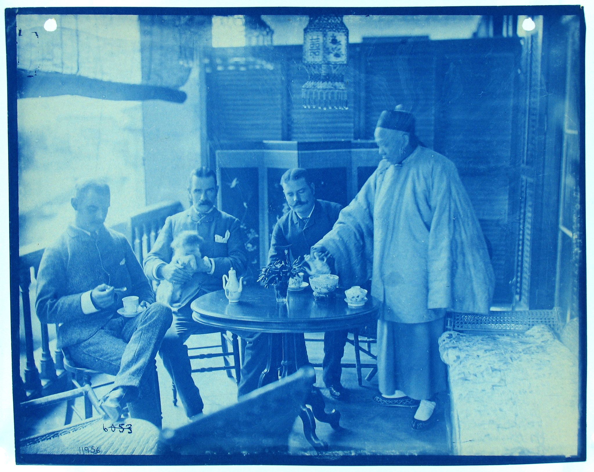 A vintage cyanotype photograph depicting a scene inside a room, likely a tea room or a domestic interior. The image is monochromatic with a distinctive blue tone characteristic of cyanotypes. The scene centers around a round table where three Western men and a Chinese man are engaged in a tea ceremony or social gathering.