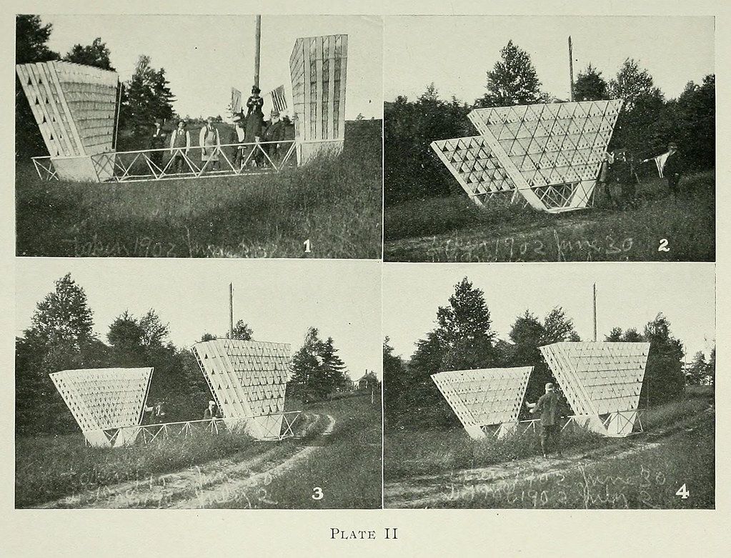 A plate with four black and white photographs arranged in a 2x2 grid. Each photograph depicts a large, triangular metal structure with a wire mesh surface, set in a grassy field with trees in the background.