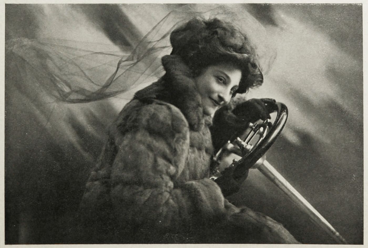 A vintage black and white photograph of a woman driving a vehicle, wearing a fur-trimmed coat and hat.