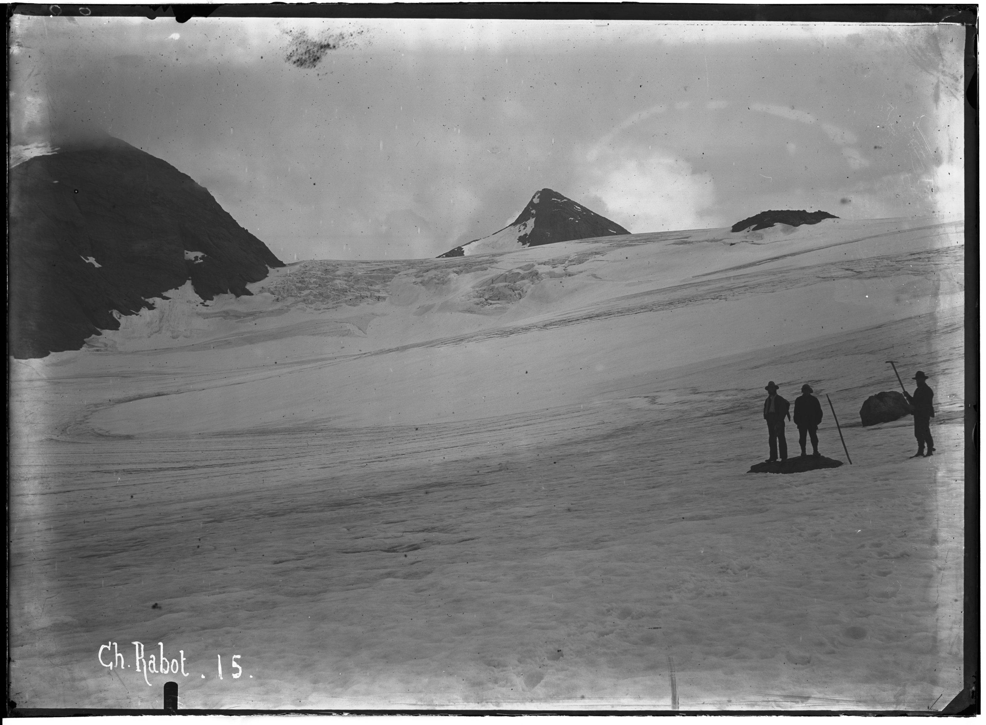 A vintage black and white photograph of a snowy landscape with three people standing on a glacier, with mountains in the background.