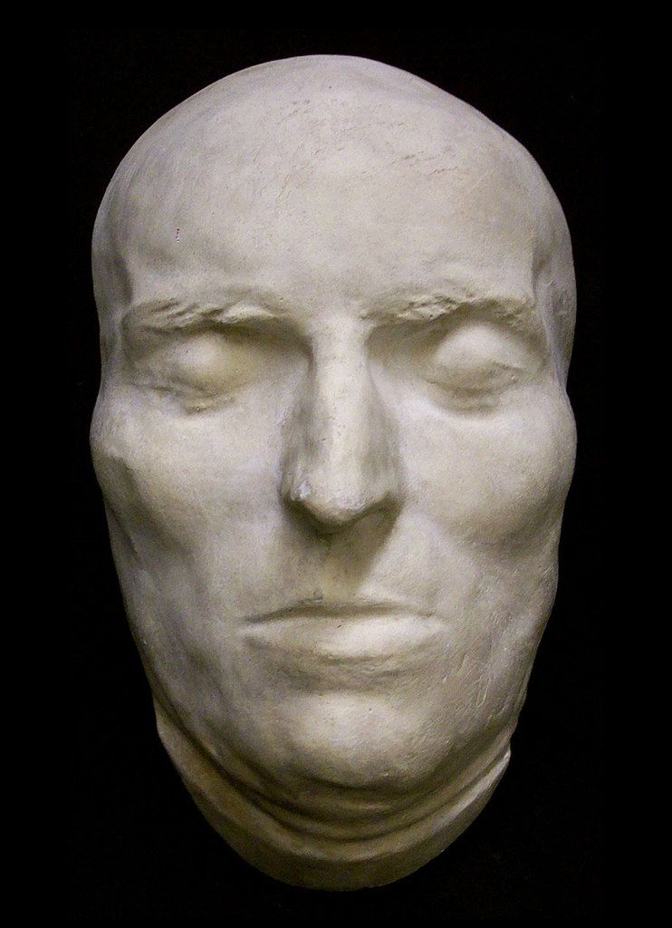 A pale plaster cast of a human face with closed eyes against a black background.