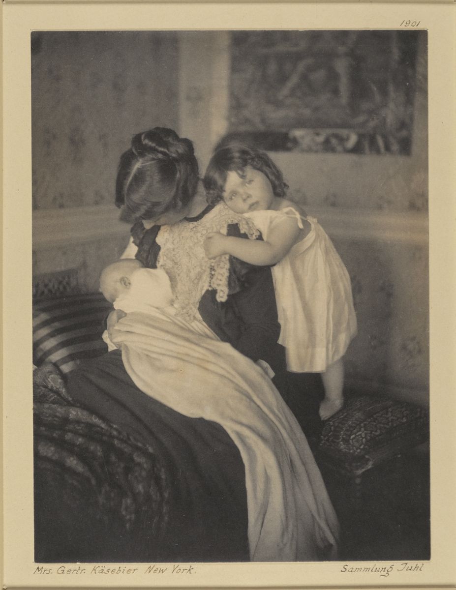 A sepia-toned photograph from 1901 depicting a woman seated on a bench holding a doll, with a young child reaching out to touch it.
