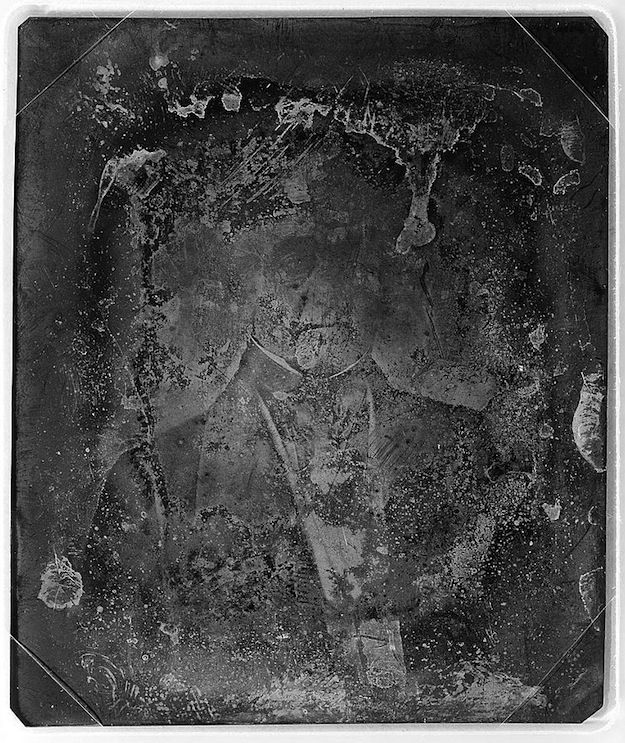 A heavily deteriorated daguerreotype showing a seated man in formal attire. The image is covered in scratches, spots, and areas of silvering loss, creating a ghostly and ethereal effect.