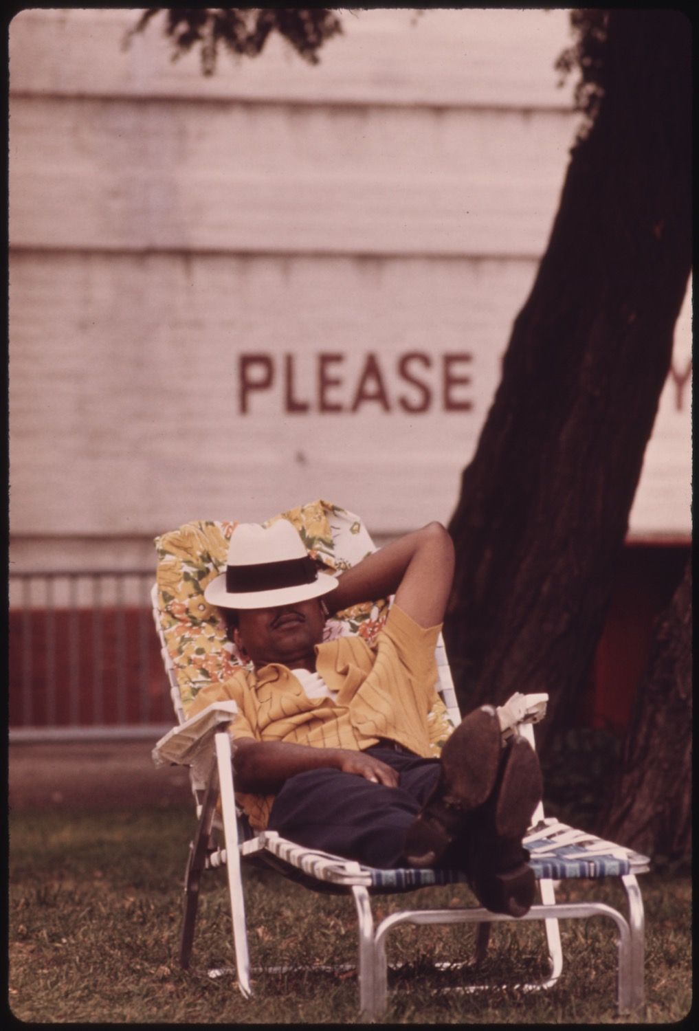 A person is relaxing in a lawn chair outdoors, wearing a white straw hat, a yellow floral shirt, and dark shorts. They are lying back with their feet propped up on another part of the chair, wearing white shoes. Behind the person is a white building with the word "PLEASE" prominently displayed in red lettering.
