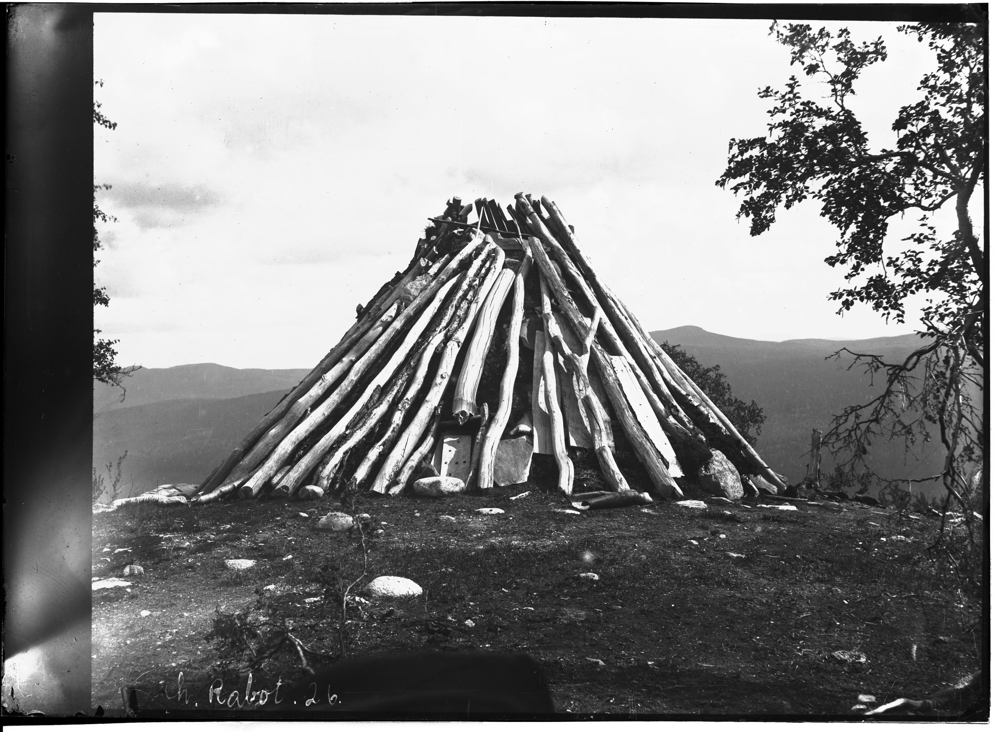 A black and white photograph of a large conical woodpile in a mountainous landscape.
