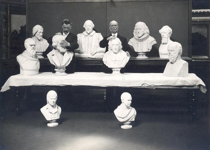 A black and white photograph of a row of plaster busts of famous historical figures displayed on a long, dark table. A man is seated between the busts, appearing to be overseeing or presenting them.