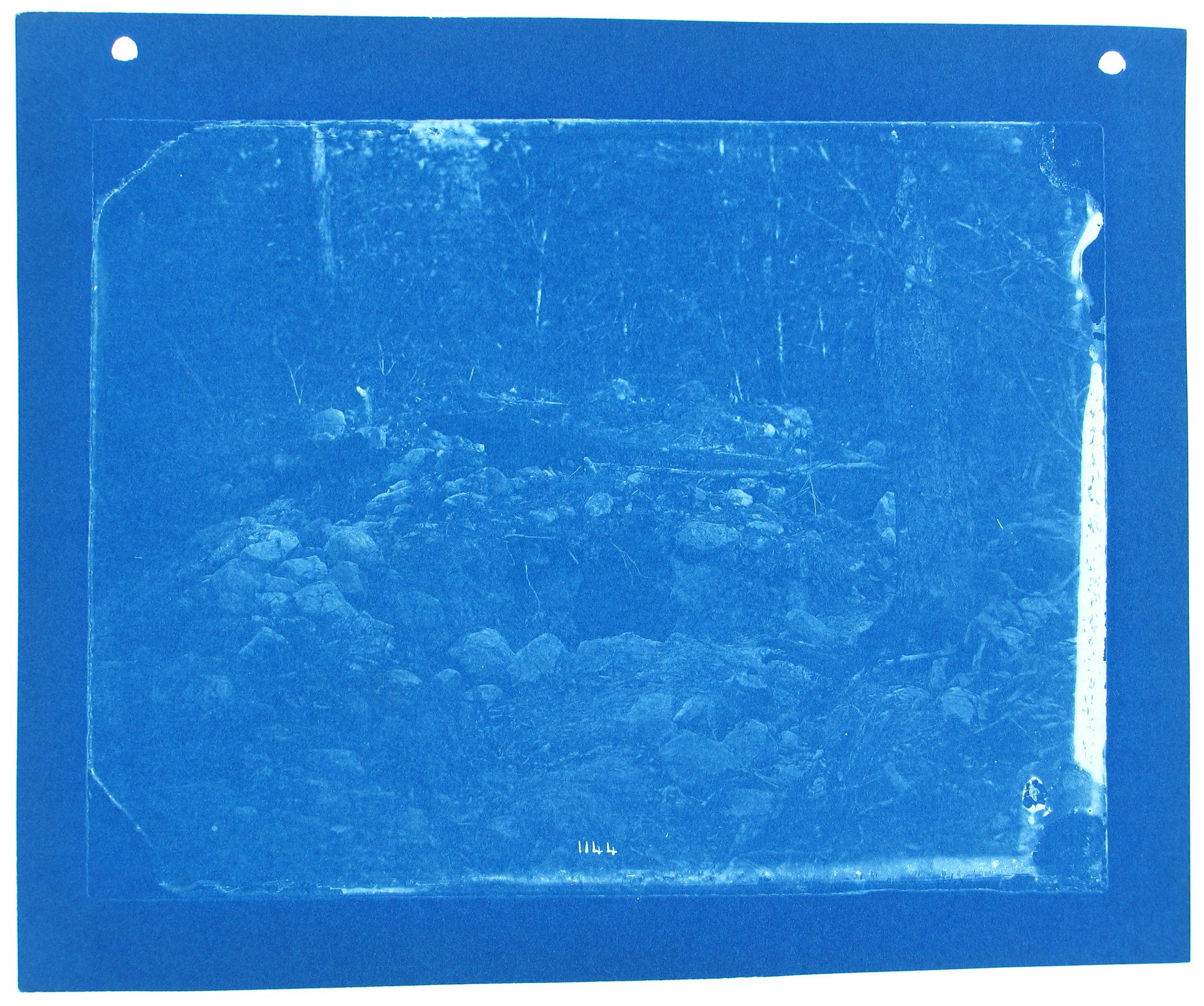 A cyanotype image of a rocky landscape with trees and a blue sky. The image is on paper with holes in the top corners and some text at the bottom.