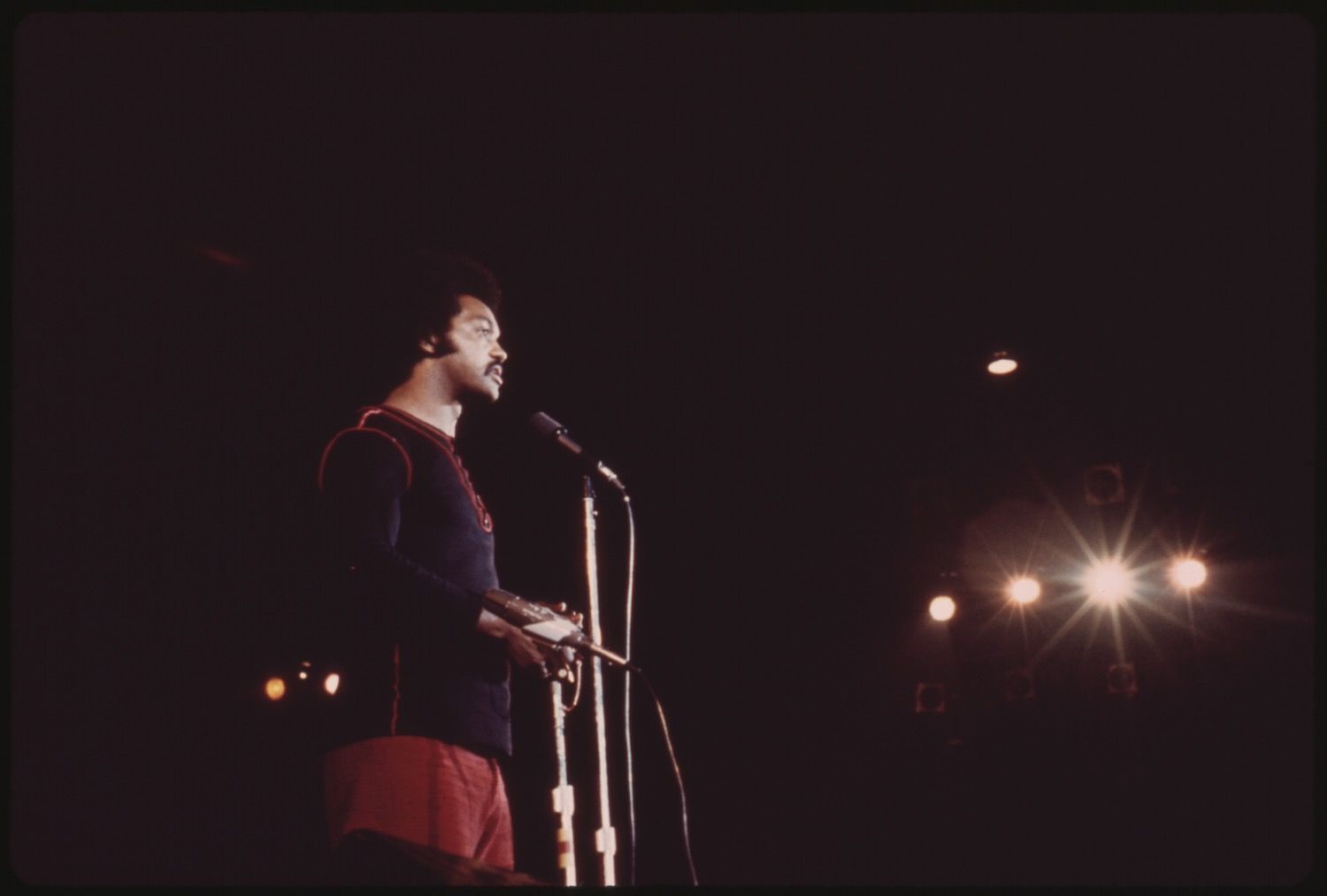 A man performing on stage with a microphone, illuminated by bright stage lights against a dark background.
