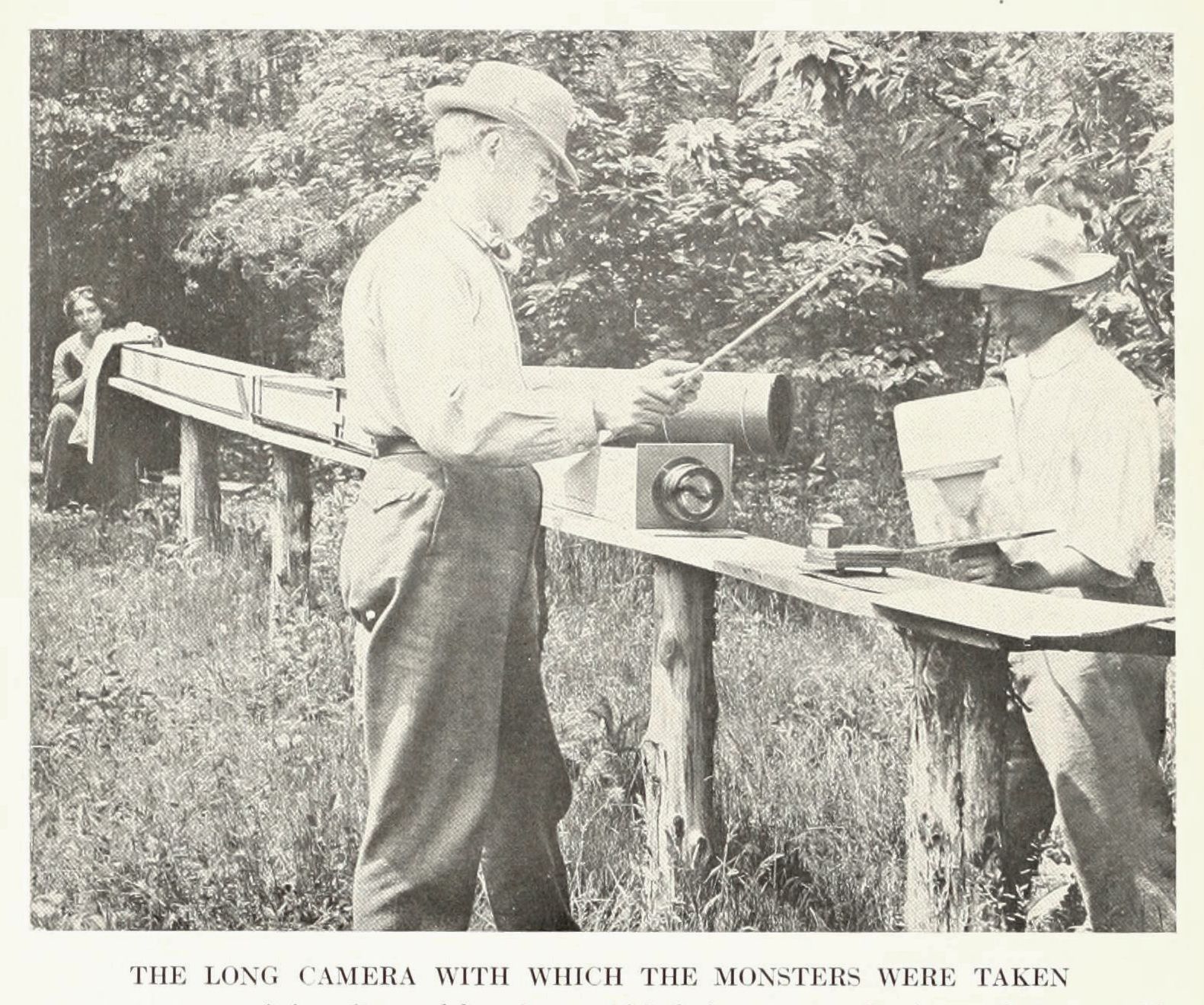 A black and white photograph of two men operating a long camera setup in a grassy, wooded area.