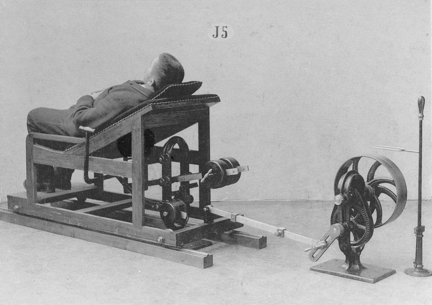 A black and white photograph of a person reclined in a complex wooden chair connected to a mechanical device.