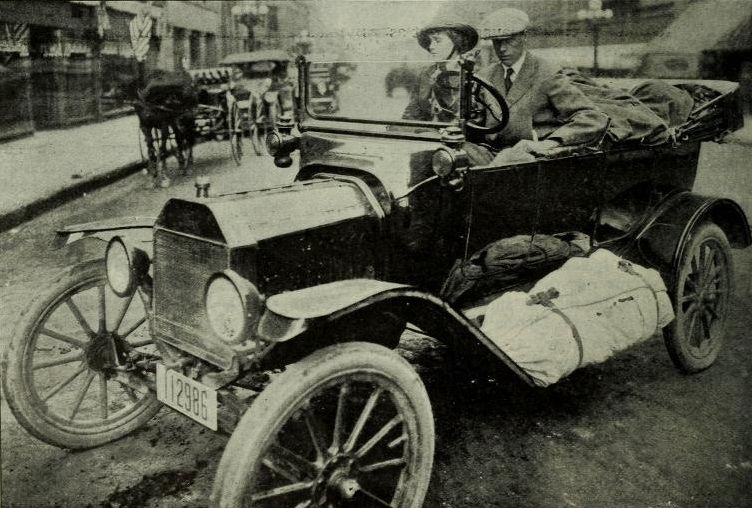 A vintage photograph of an early 20th-century automobile on a street with people and horse-drawn carriages.