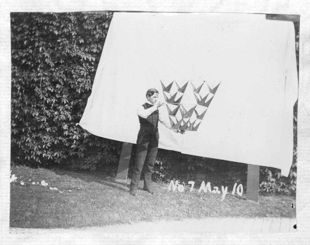 A vintage black and white photograph of a person releasing paper birds from a large white board in a grassy, tree-filled setting. The date 'No 7 May 10' is handwritten at the bottom.