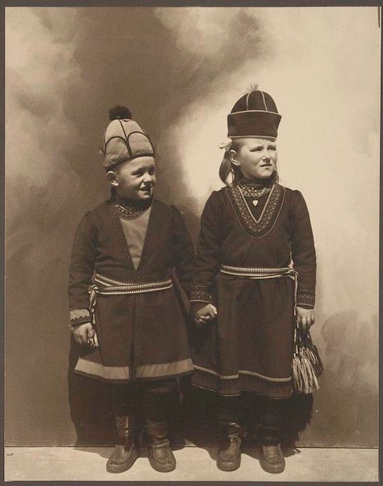 Sepia-toned photograph of two Sami women in traditional clothing, holding hands and looking at the camera.