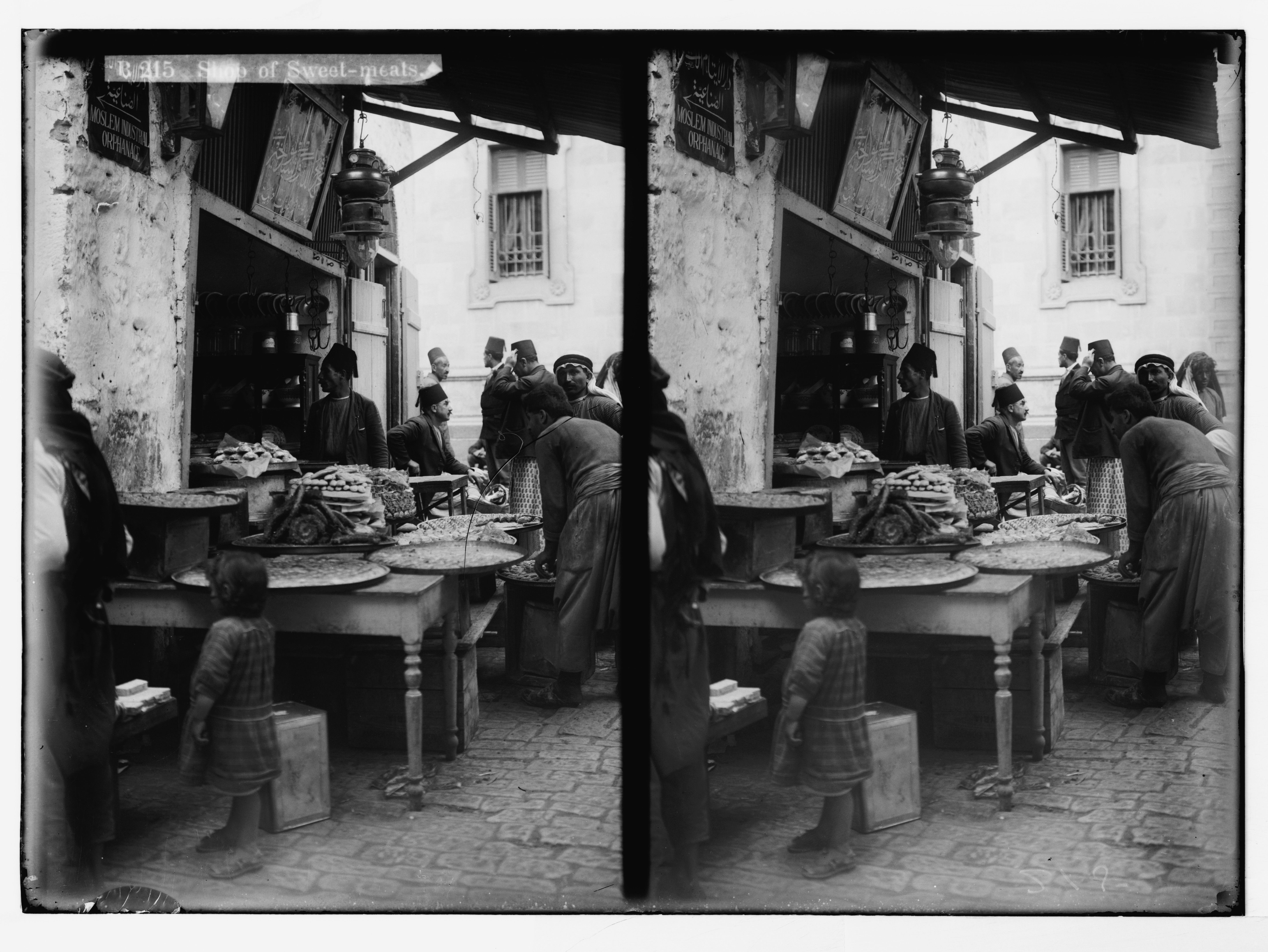 Stereoscopic photograph of a street scene with a shop selling food, people gathered around a table, and traditional clothing.