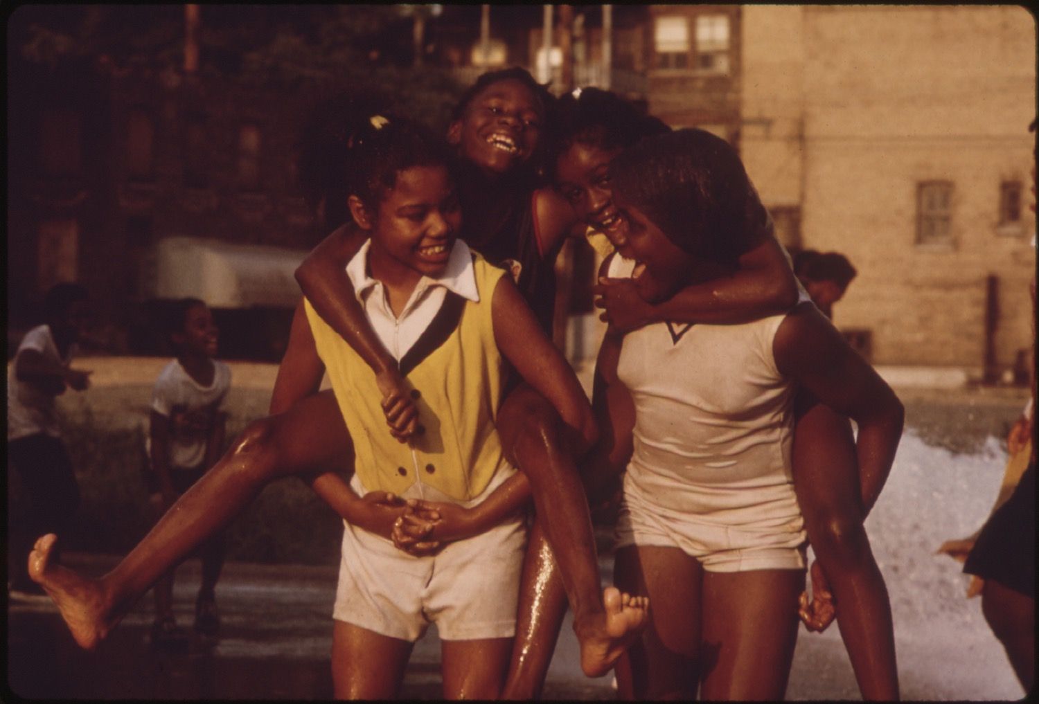 A group of three young African American girls embracing each other in a city street, wet from playing in water, with a blurred urban background.