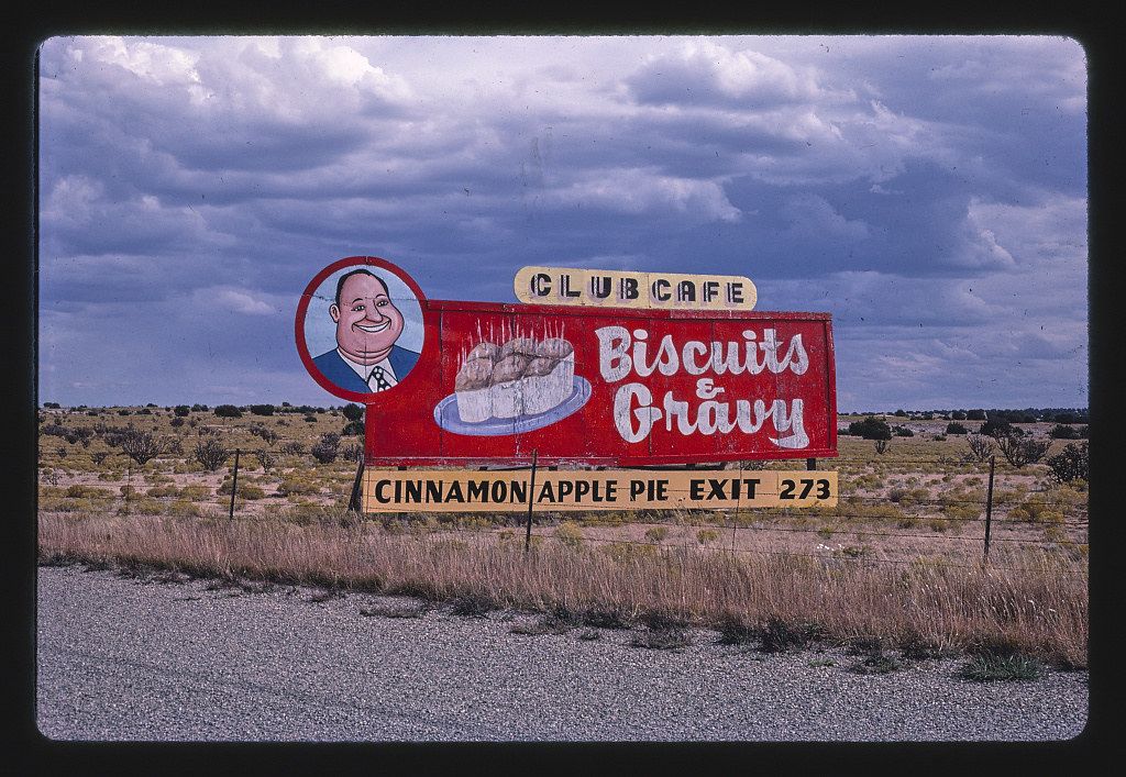 A vintage roadside sign for the "Club Cafe" advertising "Biscuits & Gravy" and "Cinnamon Apple Pie" against a backdrop of a flat, arid landscape.
