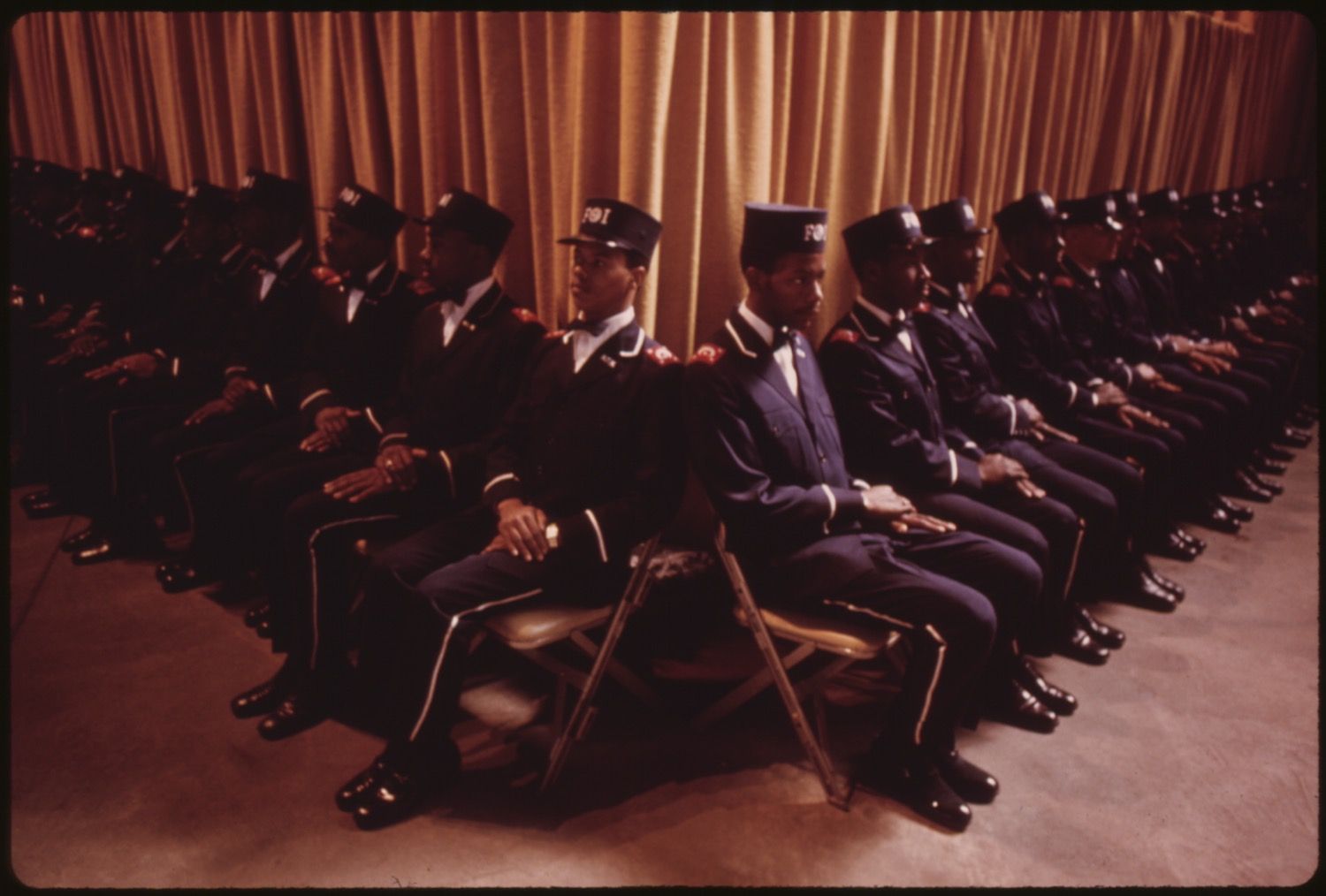 A group of African American police officers in uniform seated in rows of folding chairs in front of a golden curtain.
