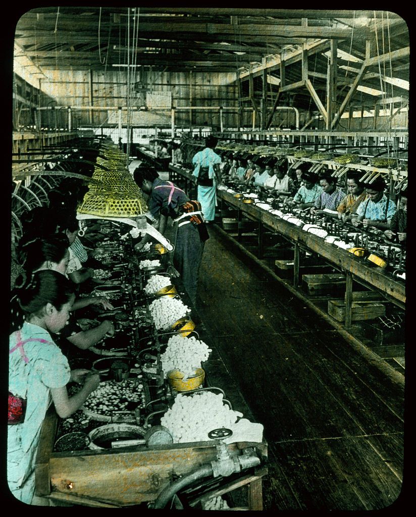 A vintage photograph of a factory interior with rows of workers at workbenches.