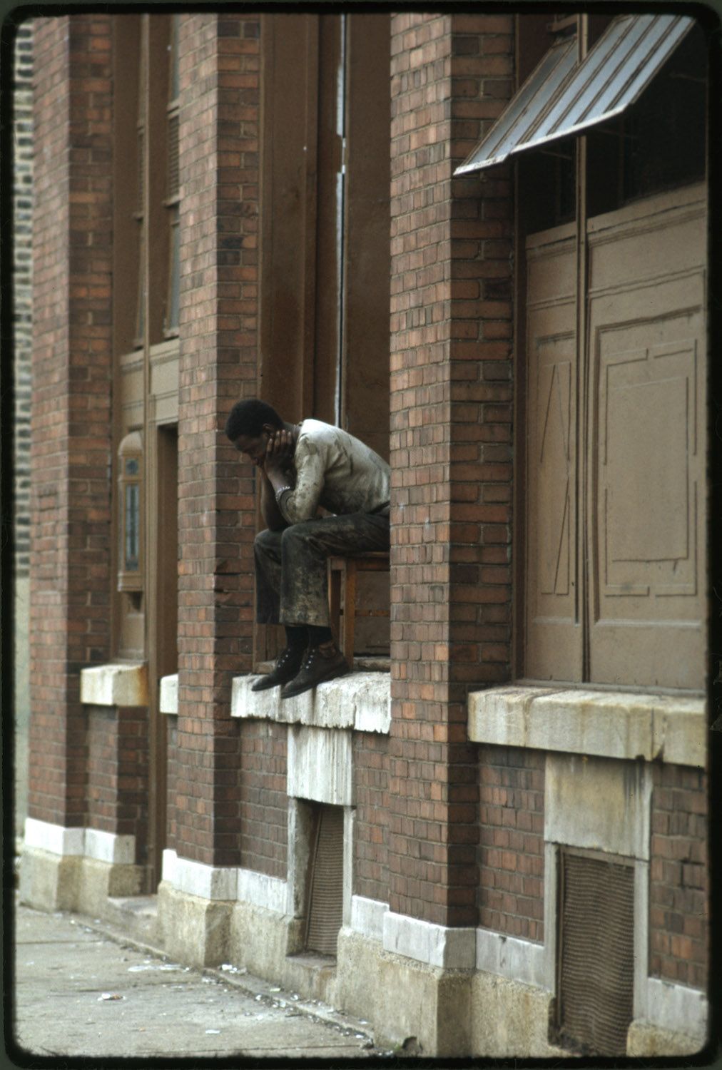 A man squats on a ledge in front of a red brick building.