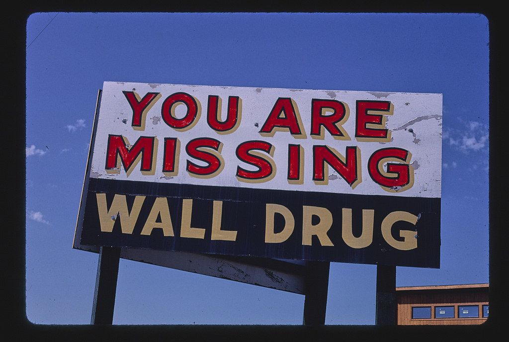 A vintage sign reading "YOU ARE MISSING WALL DRUG" against a bright blue sky.