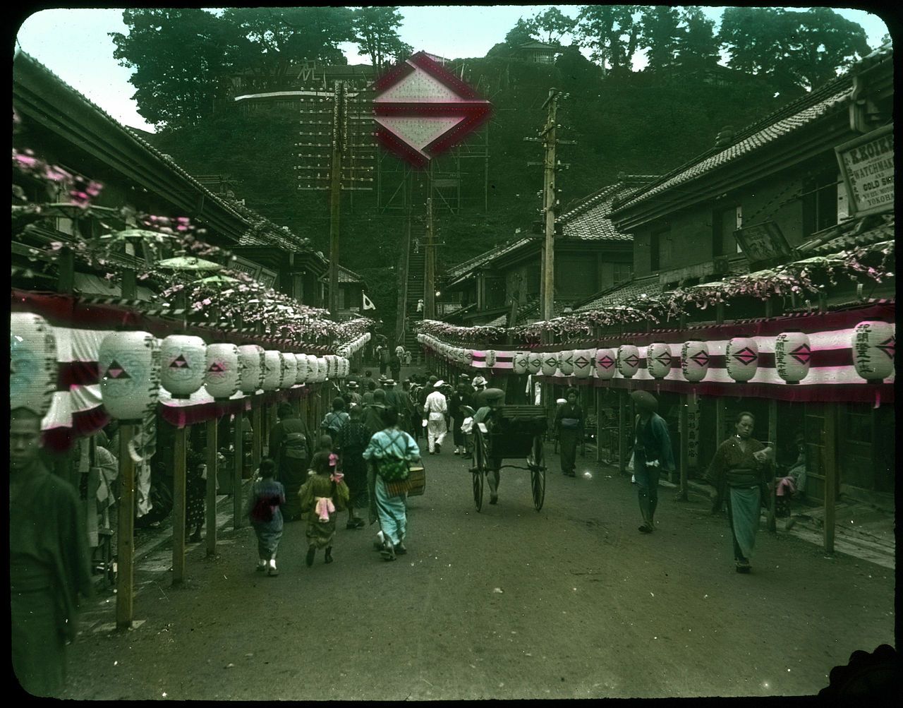 A bustling street scene in Japan, lined with traditional wooden buildings and decorated with strings of lanterns. People in a mix of traditional and Western clothing walk along the street, while a horse-drawn carriage and bicycles add to the lively atmosphere.