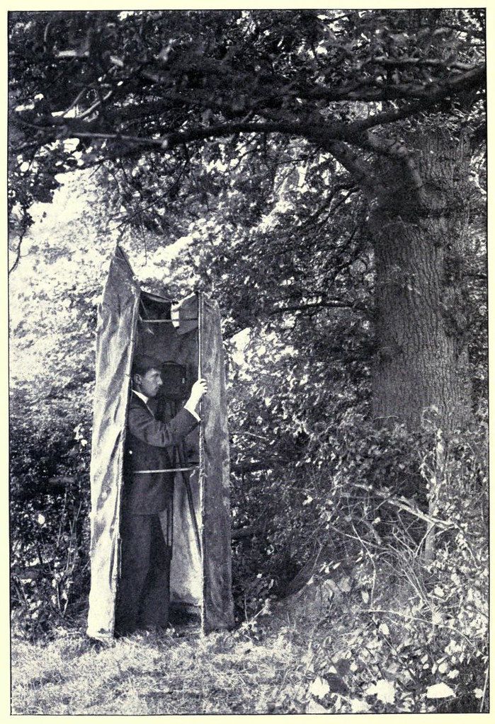 A vintage black and white photograph of a man standing inside a small, dark tent-like structure in a wooded area.
