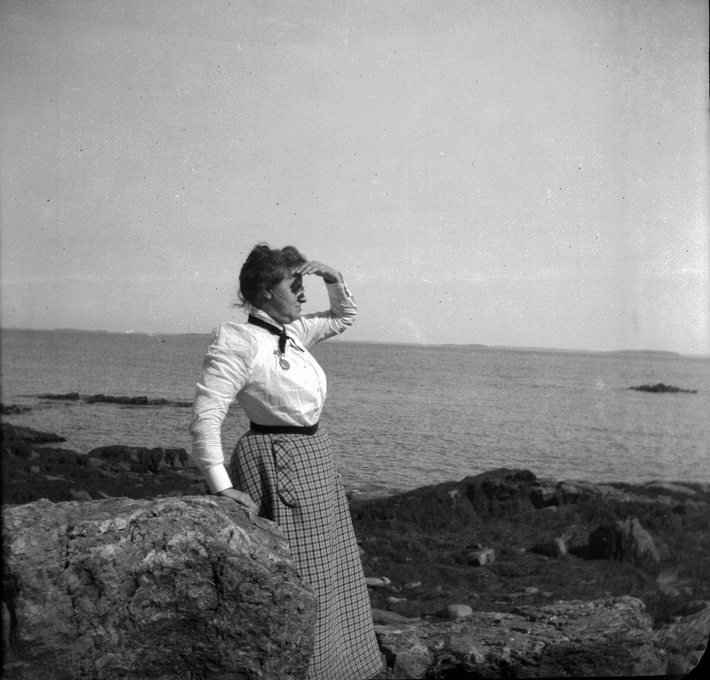 A vintage black and white photograph of a woman standing on a rocky coastline, gazing out at the sea.