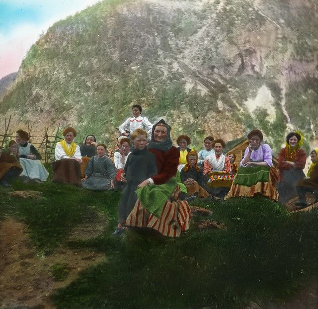 A colorized photograph of a group of people sitting on a grassy hill in front of a large, rocky hill.