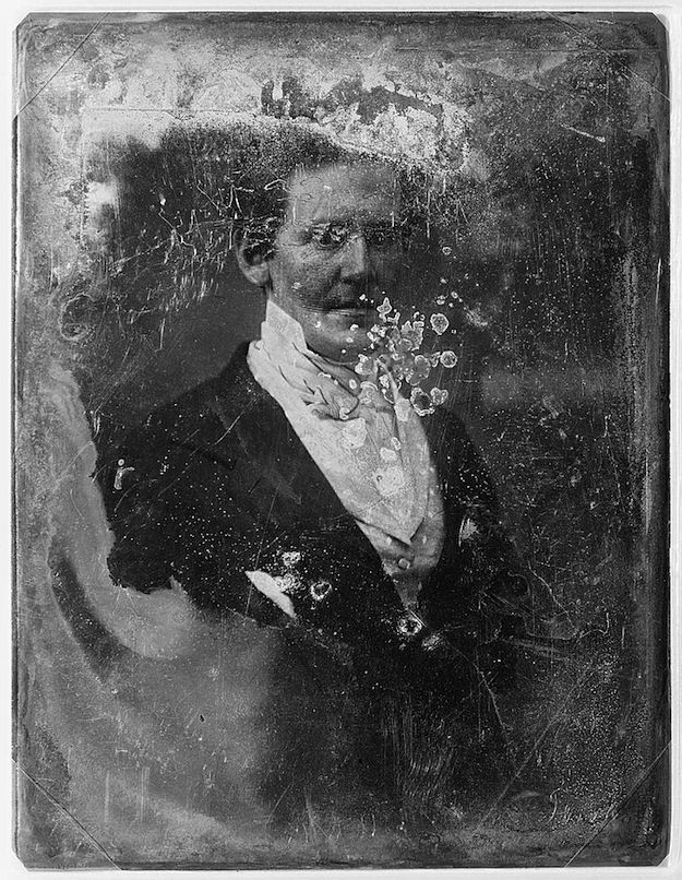 A daguerreotype portrait of a man in formal attire, with parts of his face and body disintegrating into a cloud of white particles.