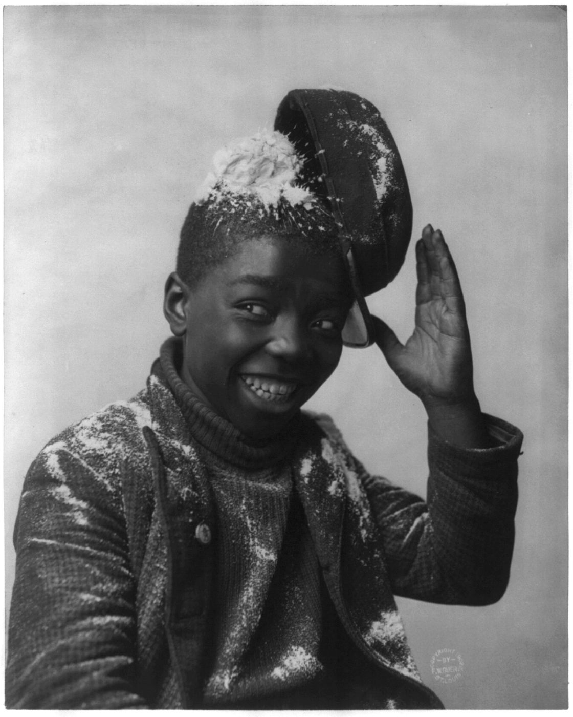 A black and white portrait of a young African American boy smiling and covered in snow, wearing a hat and coat.