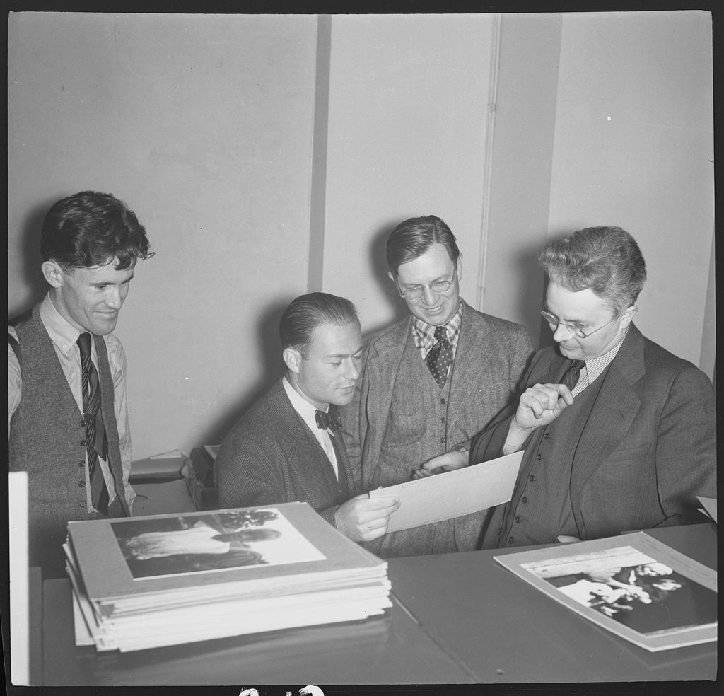 Four men in vests and ties are gathered around a table, examining a stack of photographs and a sheet of paper.