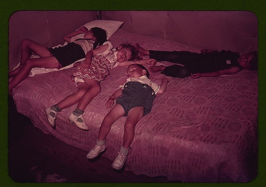 A vintage-style photograph of children lying on a patterned bed, seemingly asleep. The image has a reddish hue and is framed by a dark green border.