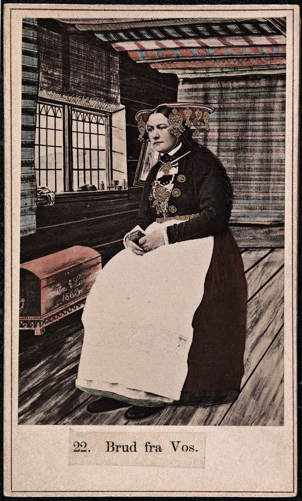 A vintage lithograph depicting a woman in traditional Norwegian folk costume, standing in a wooden interior with a window and a chest.