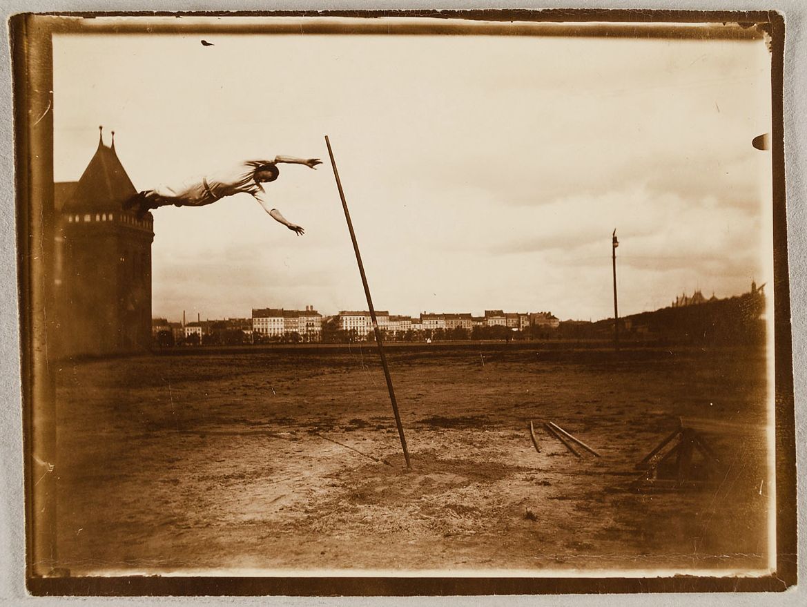 Vintage sepia-toned photograph of an athlete performing a pole vault, with a tower and cityscape in the background.