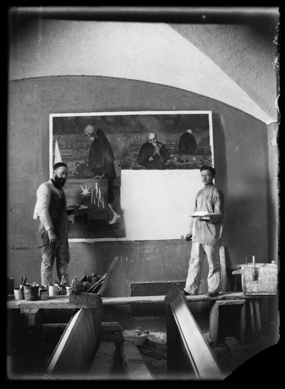 Two men stand in an artist's studio in front of a large painting on an easel.