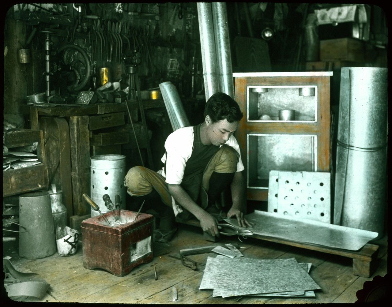 A young boy working in a cluttered workshop, surrounded by metal sheets, rolls, and tools.